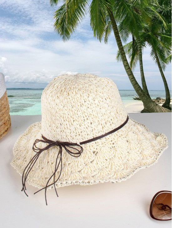 A woven white hat with brown string bow is shown on a beach backdrop.