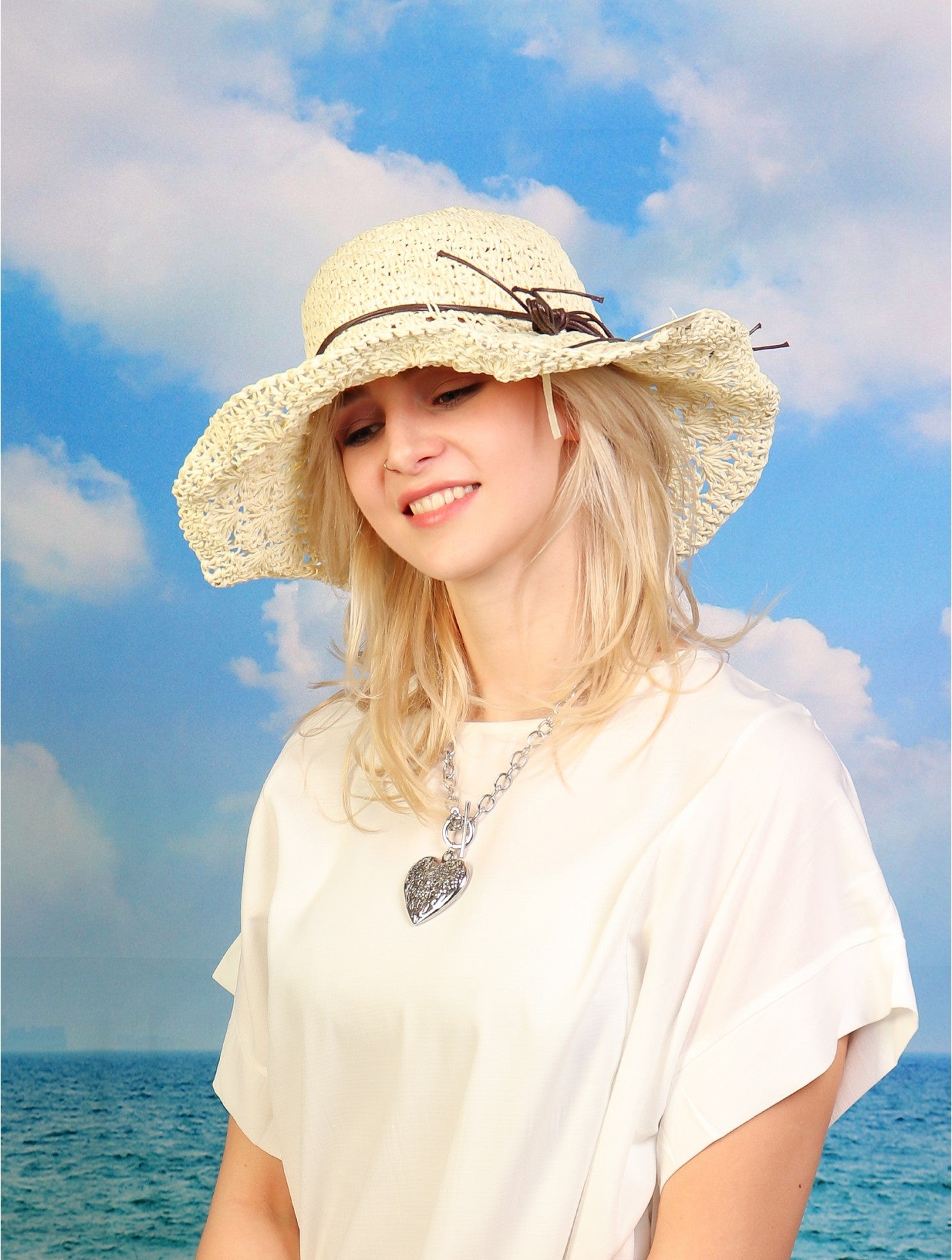 A woven white hat with brown string around the rim is shown on a woman in front of a beach backdrop.