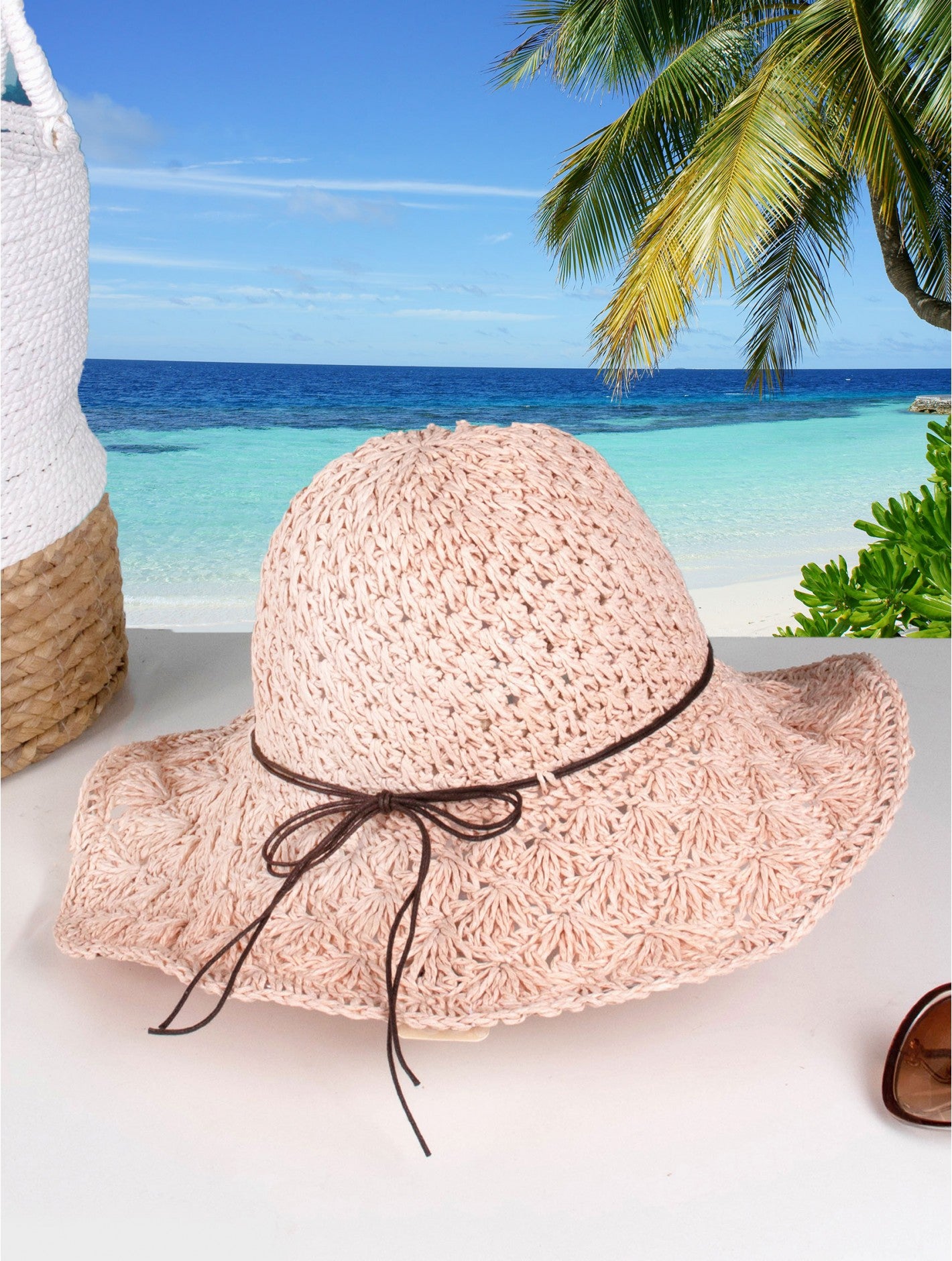 A woven pink hat with brown string bow is shown on a beach backdrop.