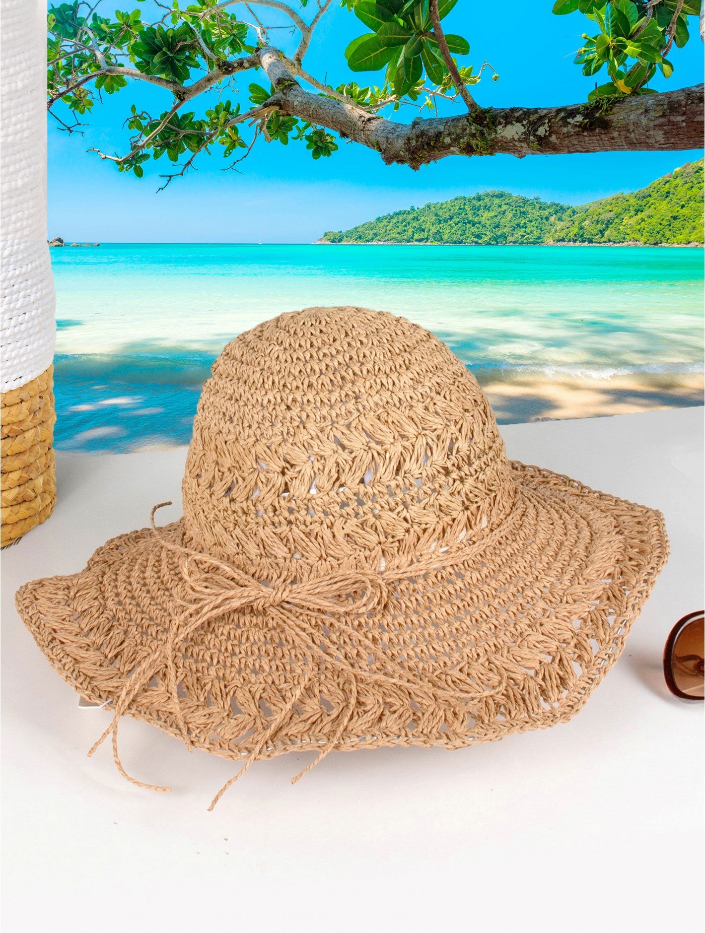 A woven light brown hat with light brown string bow is shown on a beach backdrop.